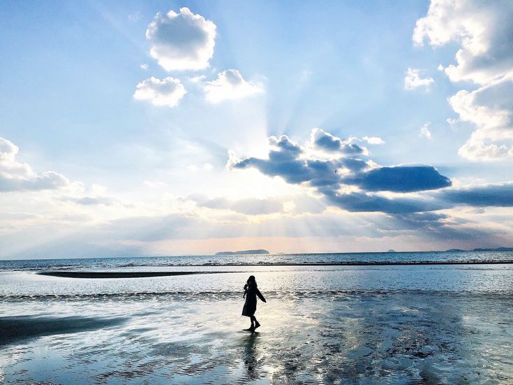 a silhouette of a person stands on a reflective beach with receding tide. the sky is filled with clouds and sun rays. the water reflects the sky and the beach stretches out to the horizon.