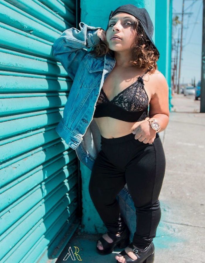 the image showcases a stylish young woman of short stature, possibly exhibiting achondroplasia,  posing confidently against a vibrant teal wall. she's dressed in a denim jacket, a black lace bra, black leggings, and highheeled shoes. her attire and pose are fashionforward, conveying a sense of selfassuredness and style. the setting is an outdoor urban environment, with the bright sunlight adding to the overall feel of the photograph.  the photo appears to be a professional photoshoot highlighting the model's personality and fashion sense.