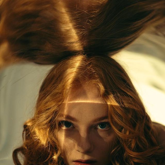 a young woman with long, curly red hair is looking at the camera. her hair is covering most of her face, but you can see her eyes, lips, and forehead. the light is coming from above and creating shadows on her face.