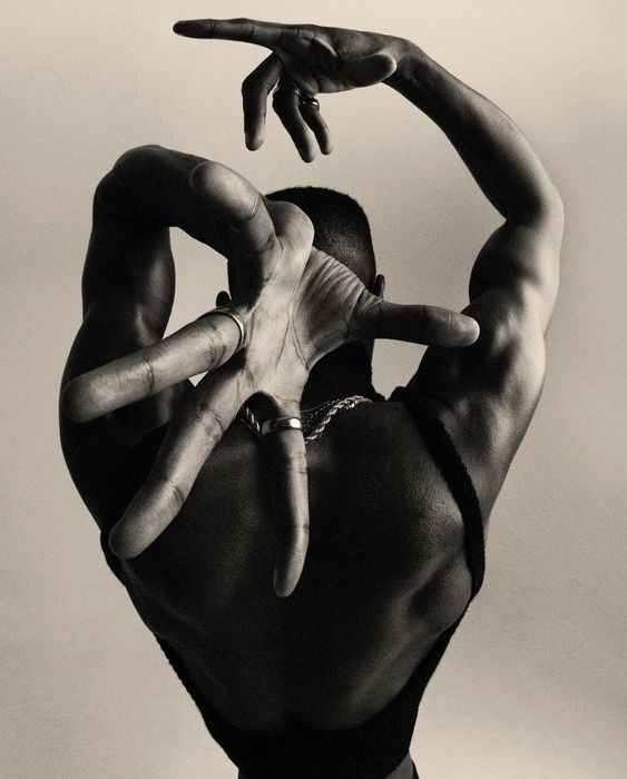 a black and white photograph of a muscular man from the back, with his arms raised and fingers intertwined in a dancelike pose. he wears rings and a chain necklace. the photo is taken in a studio with professional lighting.