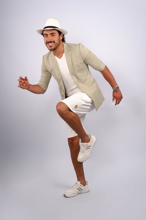 a man is posing in a studio shot. he is wearing a white hat, a beige blazer, white shorts, and white sneakers. he is also wearing a watch. he is smiling and appears to be in an active pose with one knee up.