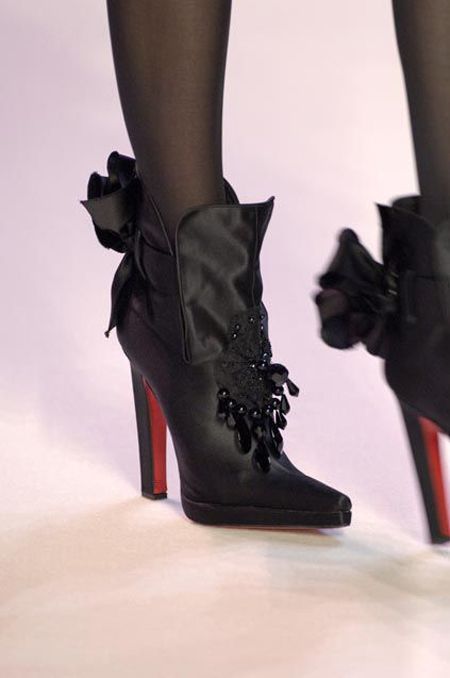 the image shows a pair of black highheeled boots adorned with bows and embellishments. the boots have a pointed toe and a red sole, indicating a luxury brand. the person wearing the boots is also wearing black tights.