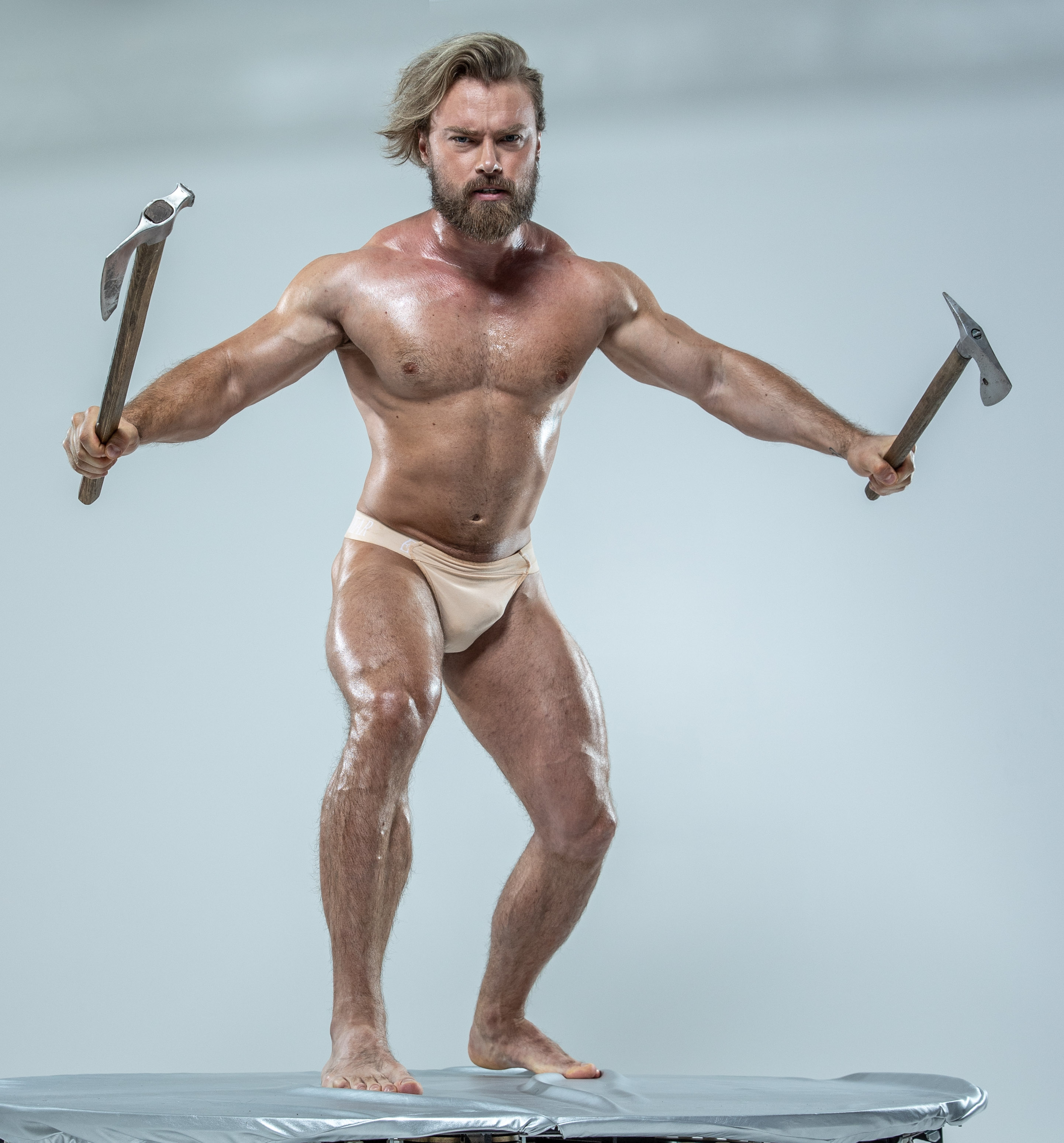 a muscular, shirtless man with styled blond hair and a beard stands with his feet planted on a round platform.  he is holding a tomahawk in each hand, and his arms are outstretched to his sides.  the background of the image is a light gray.