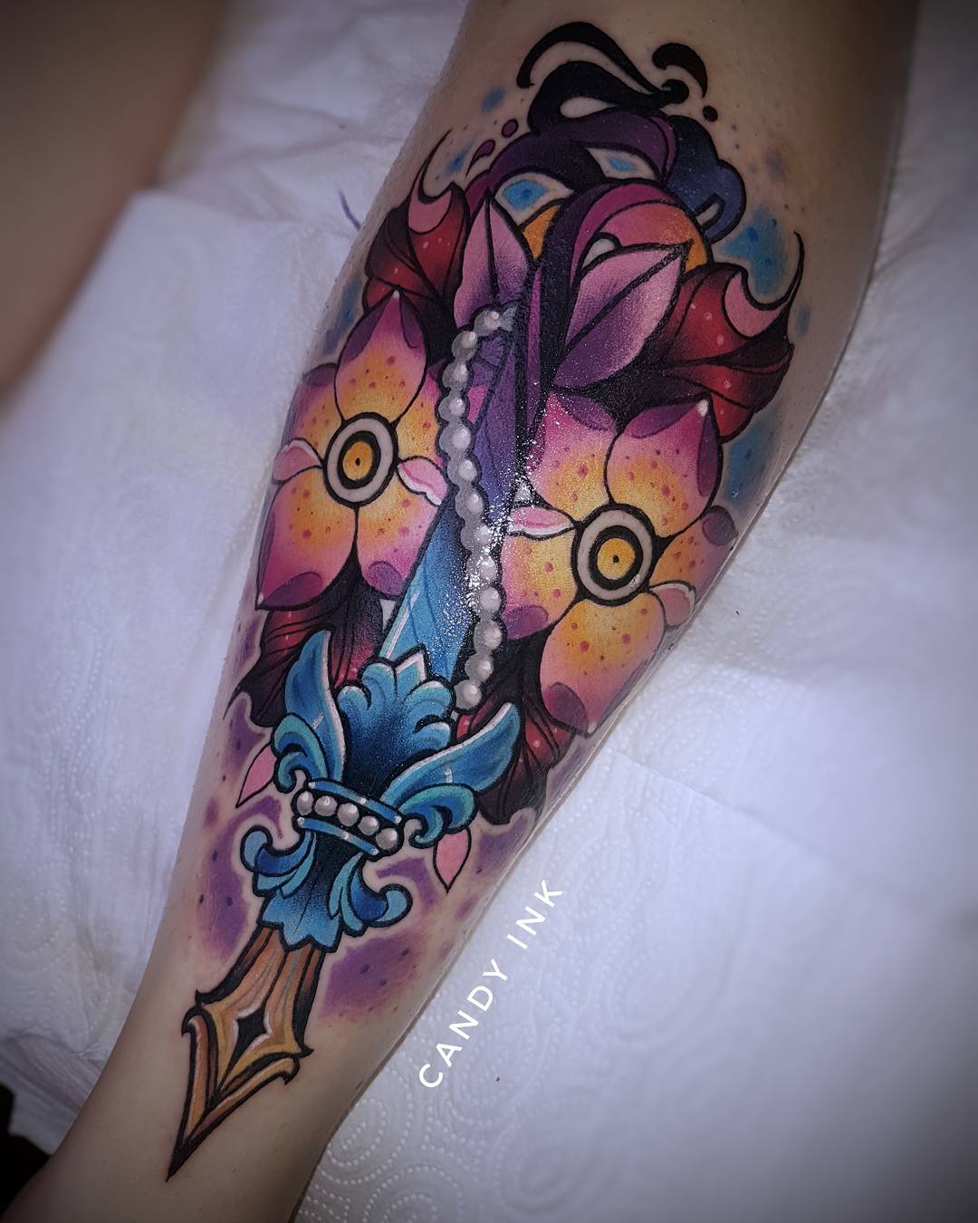 a person's leg with a colorful neotraditional tattoo of a dagger with a fleurdelis pommel and flowers. the tattoo is done in a bright and bold style with thick black outlines.