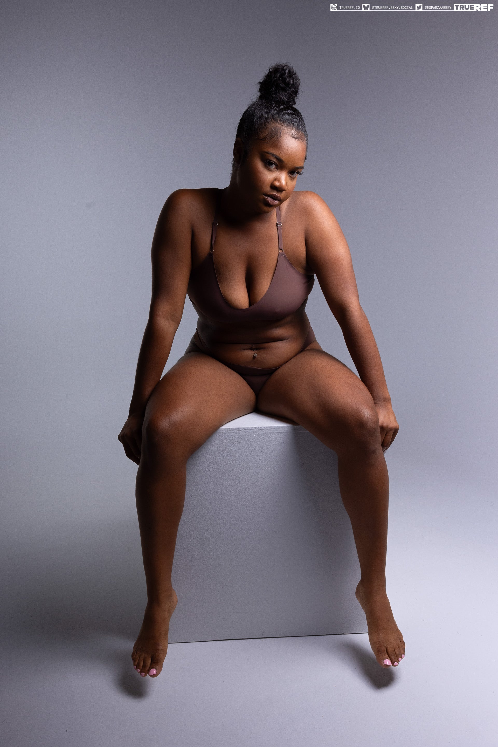 a medium shot studio portrait of a confident plussize woman with dark skin sitting on a white cube against a gray background. she is wearing brown lingerie. her hair is in a bun. she is barefoot with nail polish on her toes. she is looking directly at the camera with a confident gaze.