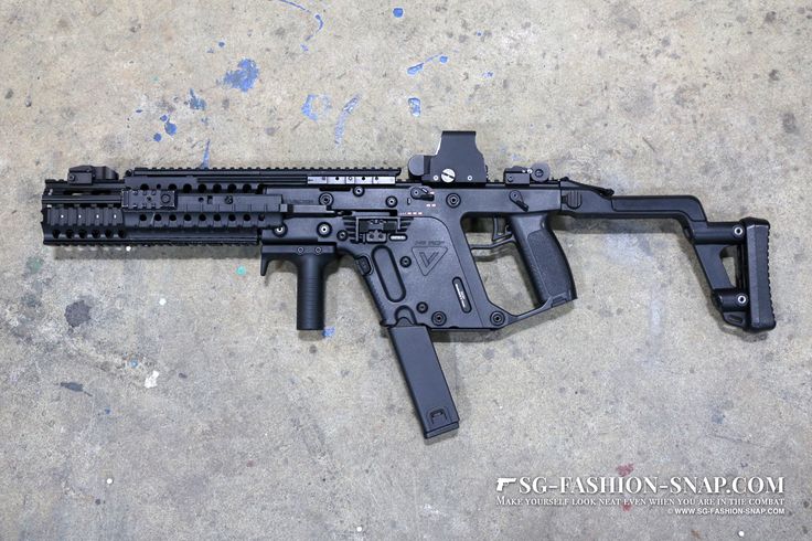 a black kriss vector submachine gun is lying on a gray concrete surface. the weapon has a foregrip, optic, magazine, and stock. it appears to be a compact and tactical firearm.