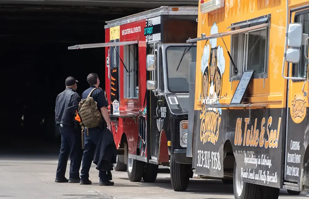 two food trucks are parked sidebyside, one red and one yellow.  a pair of men are standing in front of the trucks, apparently deciding which one to order from.  the red food truck has a sign that says "we cater all events" and "detroit food."  the yellow food truck has a sign that says "oh taste & see!" and "specializing in catering, private events and daily specials."