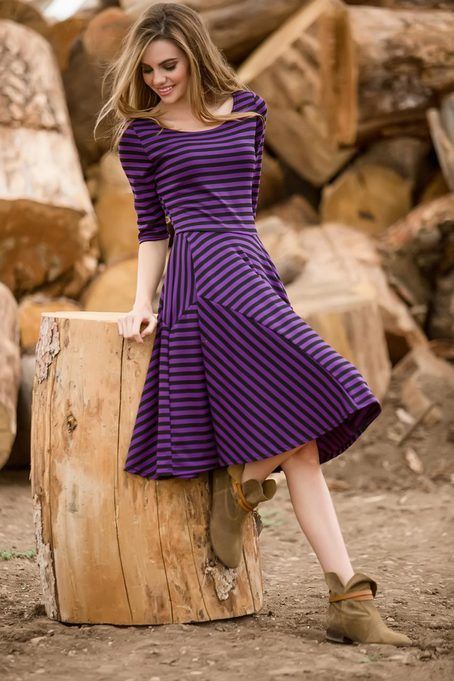 a young woman with long brown hair, dressed in a purple and black striped dress, stands beside a large log outdoors. she is smiling and looking down at the camera. she is wearing brown boots.