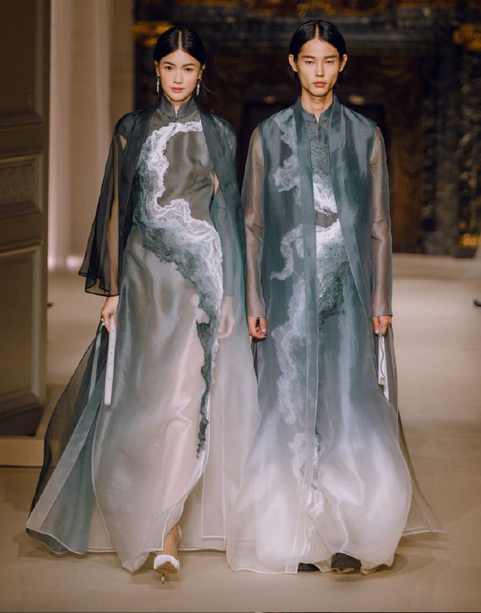 two asian models, a woman and a man, are walking on a runway during a fashion show. they are both wearing traditionalinspired clothing made of sheer fabric with a gradient effect and intricate embroidery details. the woman is wearing a dress and a sheer outer layer, while the man is wearing a matching outfit. the clothing is elegant and stylish, with long sleeves and a high collar. the background is light and softly lit, emphasizing the clothing and the models.