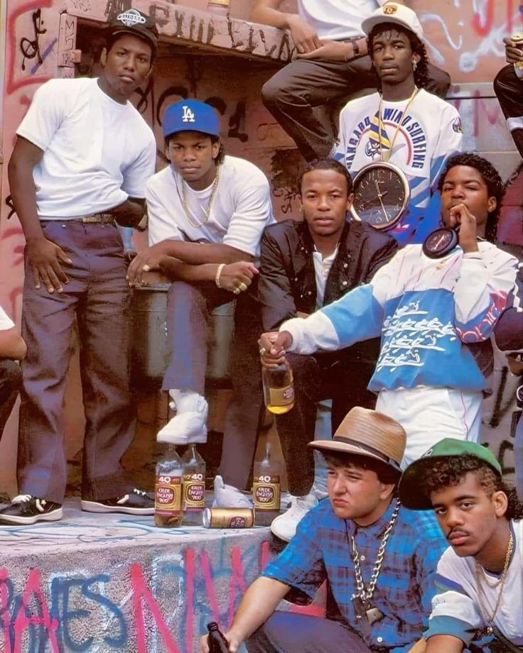 the image is a vintage photograph depicting the iconic hiphop group n.w.a.  all members are present: ice cube, eazye, dr. dre, mc ren, and dj yella. they are posed casually in an urban setting against a graffiticovered wall. the members are wearing 1980sstyle clothing and accessories, including baseball caps and oversized shirts.  several 40 ounce bottles of beer are visible on the ground in the foreground, contributing to the group's image. the overall vibe is one of raw energy and street authenticity, reflecting the genre and era. the photo captures a candid moment of the group, conveying their attitude and style.