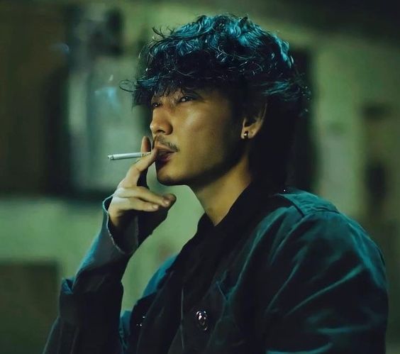 a young man with tousled dark hair and a mustache is captured in a closeup, smoking a cigarette. his expression is contemplative and slightly forlorn. the setting appears to be an urban environment, likely indoors, with a dimly lit atmosphere. the lowkey lighting accentuates the moodiness of the scene, while the shallow depth of field focuses attention on the subject. the man's attire, consisting of a dark jacket and shirt, further reinforces the overall edgy and grunge aesthetic. the image evokes a cinematic quality, reminiscent of a still from a film.