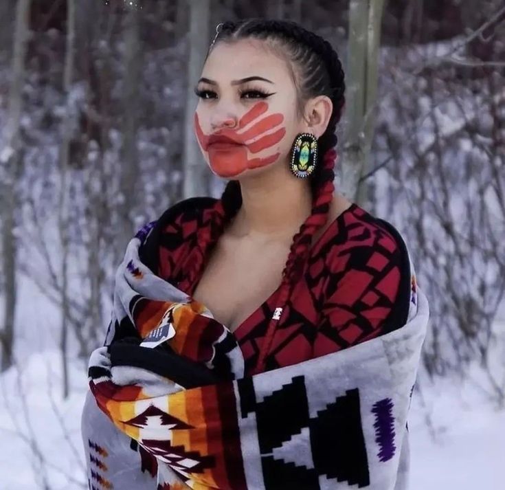 a native american woman with her dark hair in two braids, wears a traditional patterned blanket while standing in a snowy outdoor setting. her face is painted with red handprints.  she is looking directly at the camera.