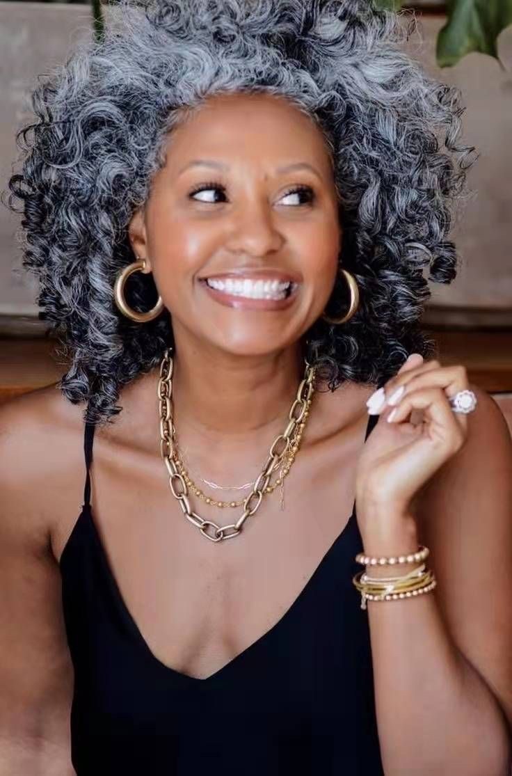 a portrait of a beautiful woman with grey hair smiling, wearing a black top and gold jewelry. she looks happy and confident. the woman is wearing large gold hoop earrings, a layered necklace with large gold chain links, and gold bracelets on her wrist. her grey hair is styled in loose curls. she has a natural look and appears to be in her midthirties to midforties. the background is out of focus but appears to be a home or office setting.