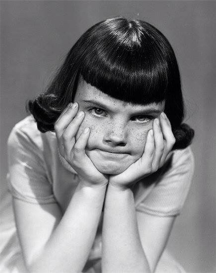 a black and white portrait of a young girl with a serious expression. she is resting her chin on her hands and looking down with a bored and frustrated expression.