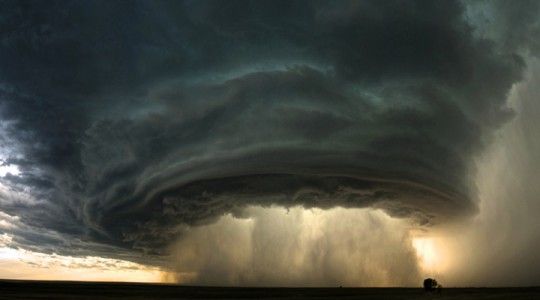a stunning image of a massive dark storm cloud with a funnel cloud rotating at the bottom. the storm is taking place over a landscape that appears to be a field with a lone tree in the distance. the dark clouds fill the majority of the image, and the sun is setting or rising in the distance, creating a glowing effect behind the storm. the image is a powerful and dramatic depiction of the raw power of nature.