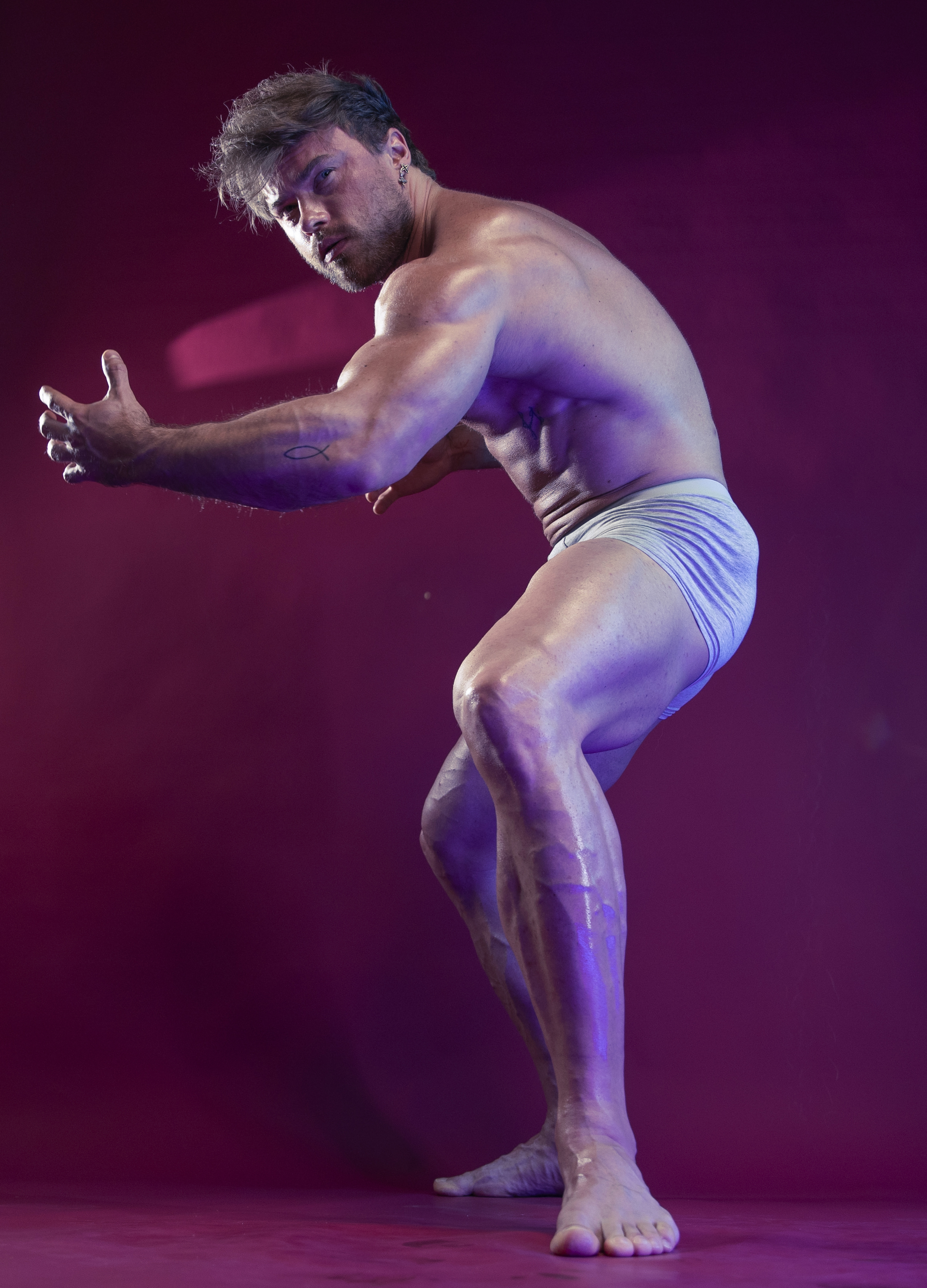 a muscular, shirtless man in briefs poses in a studio against a pink background. he is facing left, with his body angled slightly towards the viewer. his arms are bent at the elbows, with his hands open. he has a tattoo on his left arm. the lighting highlights his musculature and veins.
