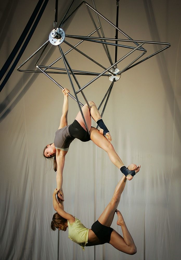 two women are performing an aerial silks routine, suspended high in the air, one hanging upside down while the other holds her upside down and poses on her legs.