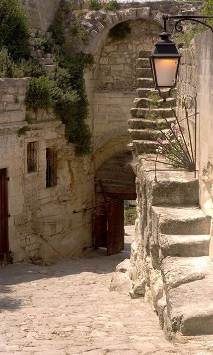 this image captures a picturesque old town scene with a narrow alleyway featuring stone steps and a vintage street lamp. the scene is dominated by aged stone buildings, an archway leading further into the town, a doorway, and a cobblestone pavement. various plants add a touch of nature to the architectural scene.