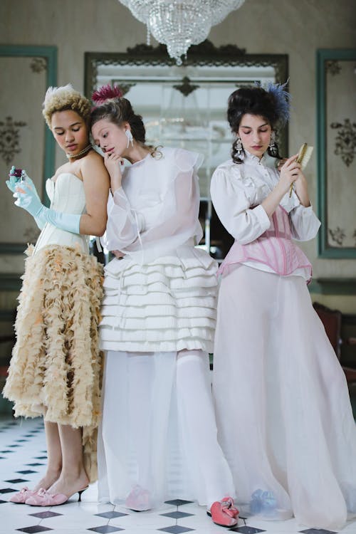 hree women in elegant, vintagestyle dresses pose in an antique setting. the women are dressed in pastelcolored gowns with elaborate details like lace, ruffles, and feather accents. they hold various objects, suggesting a scene from a period drama or historical reenactment. the background features an antique mirror reflecting the ornate interior of the room. the overall aesthetic is refined, conveying a sense of history and elegance.