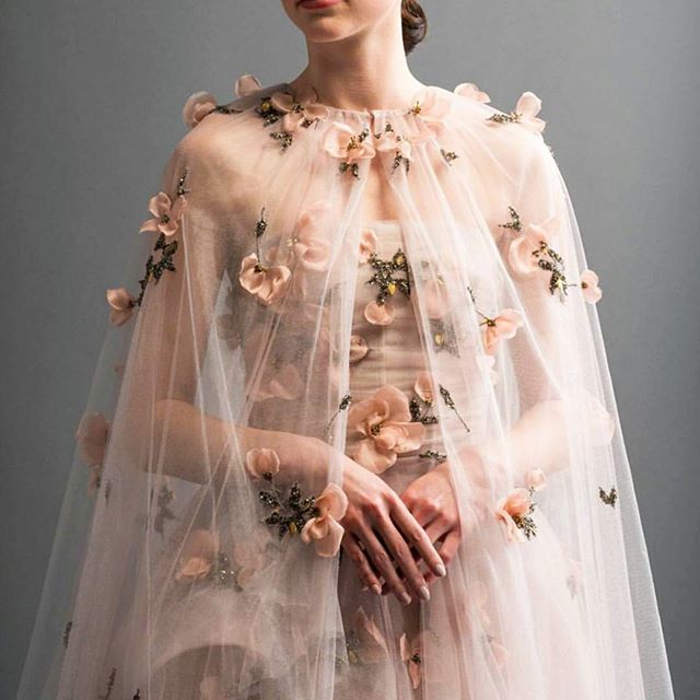 a model is wearing a peachcolored formal gown with a sheer, light pink tulle cape adorned with peachcolored flowers and beaded details. the cape drapes over the shoulders and extends down to the floor. the model's hands are clasped in front, showcasing the delicate details of the dress and cape. the background is a neutral gray.