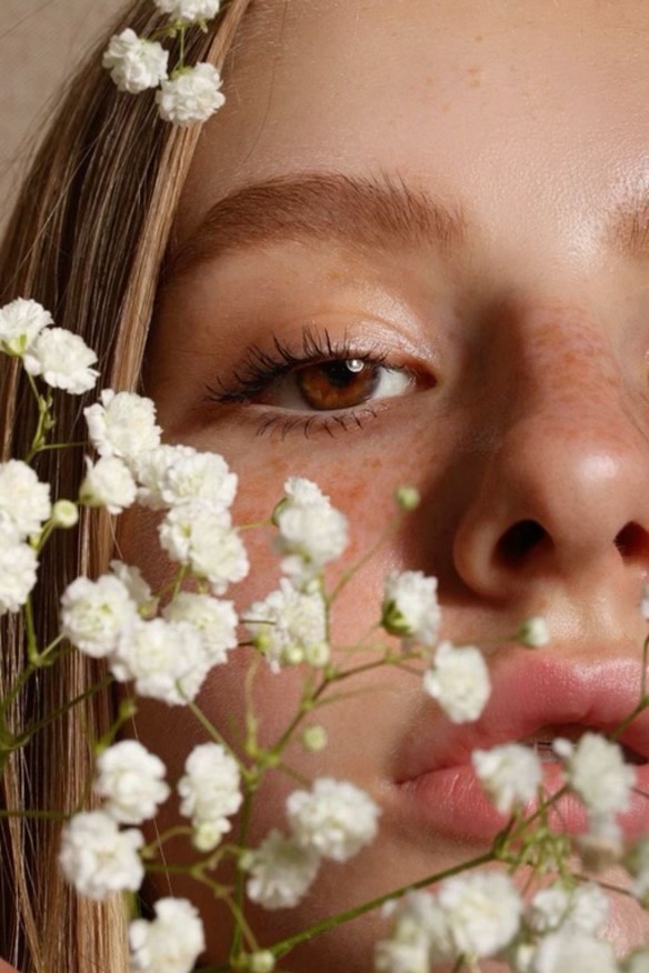 a closeup portrait of a young woman's face partially obscured by a bouquet of white baby's breath flowers.  the woman has light skin, freckles, blonde hair, and brown eyes with soft, naturallooking makeup. the overall aesthetic is delicate, romantic, and peaceful.  the image is welllit, emphasizing the soft textures of the skin and flowers.