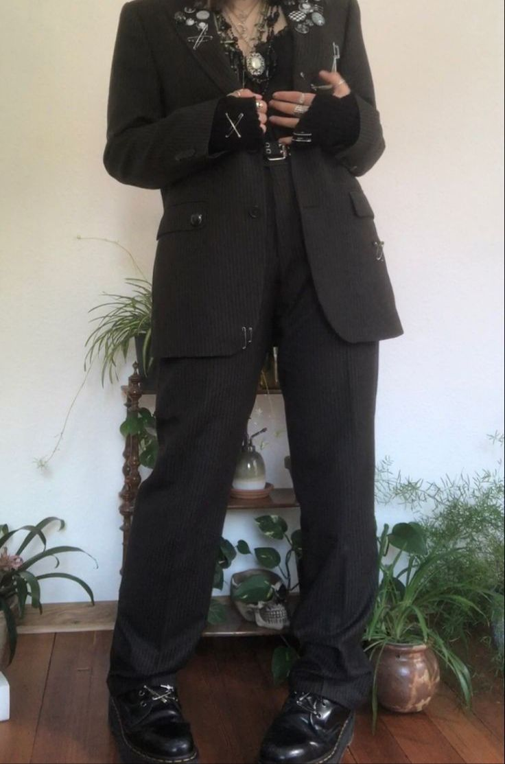 a person wearing a black pinstriped blazer and pants, with black boots and several accessories including silver chains, pins, rings, and gloves. the person is standing in front of a wooden shelf with plants.  the photo is taken from a low angle, focusing on the clothing and accessories.