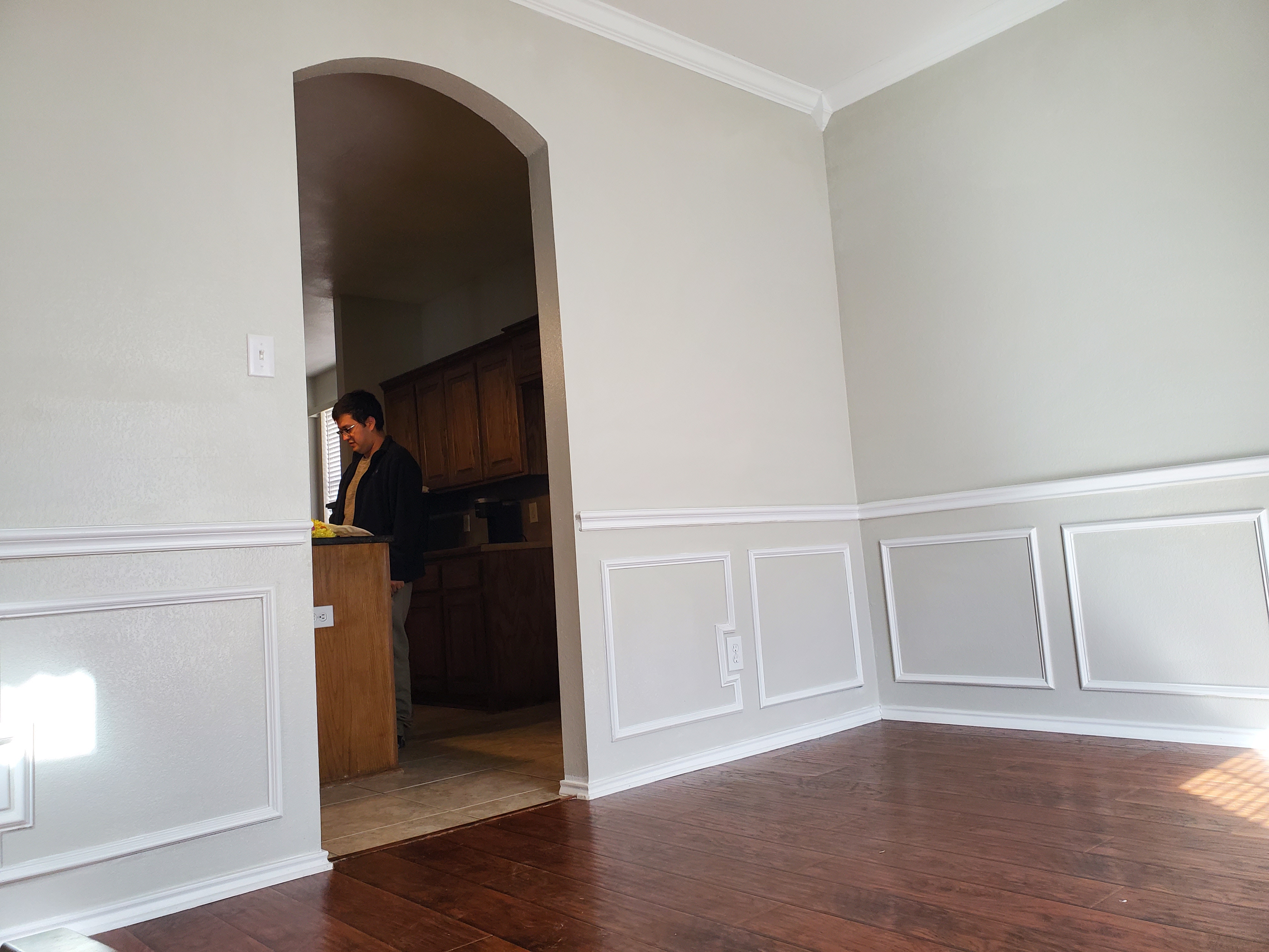 the photo shows the interior of a room with a dark hardwood floor, light grey walls with white molding and wainscoting. an archway leads to a kitchen with wooden cabinets. a man is partially visible in the kitchen. the dining room is empty and well lit.