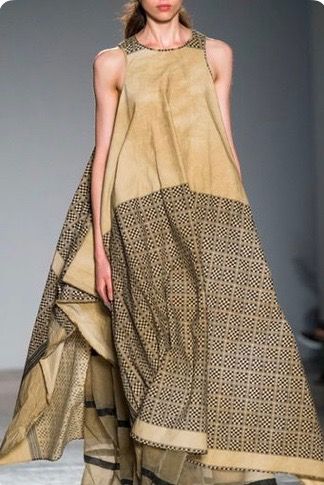 a woman is walking on a runway wearing a long, beige sleeveless dress with a patterned fabric at the bottom.