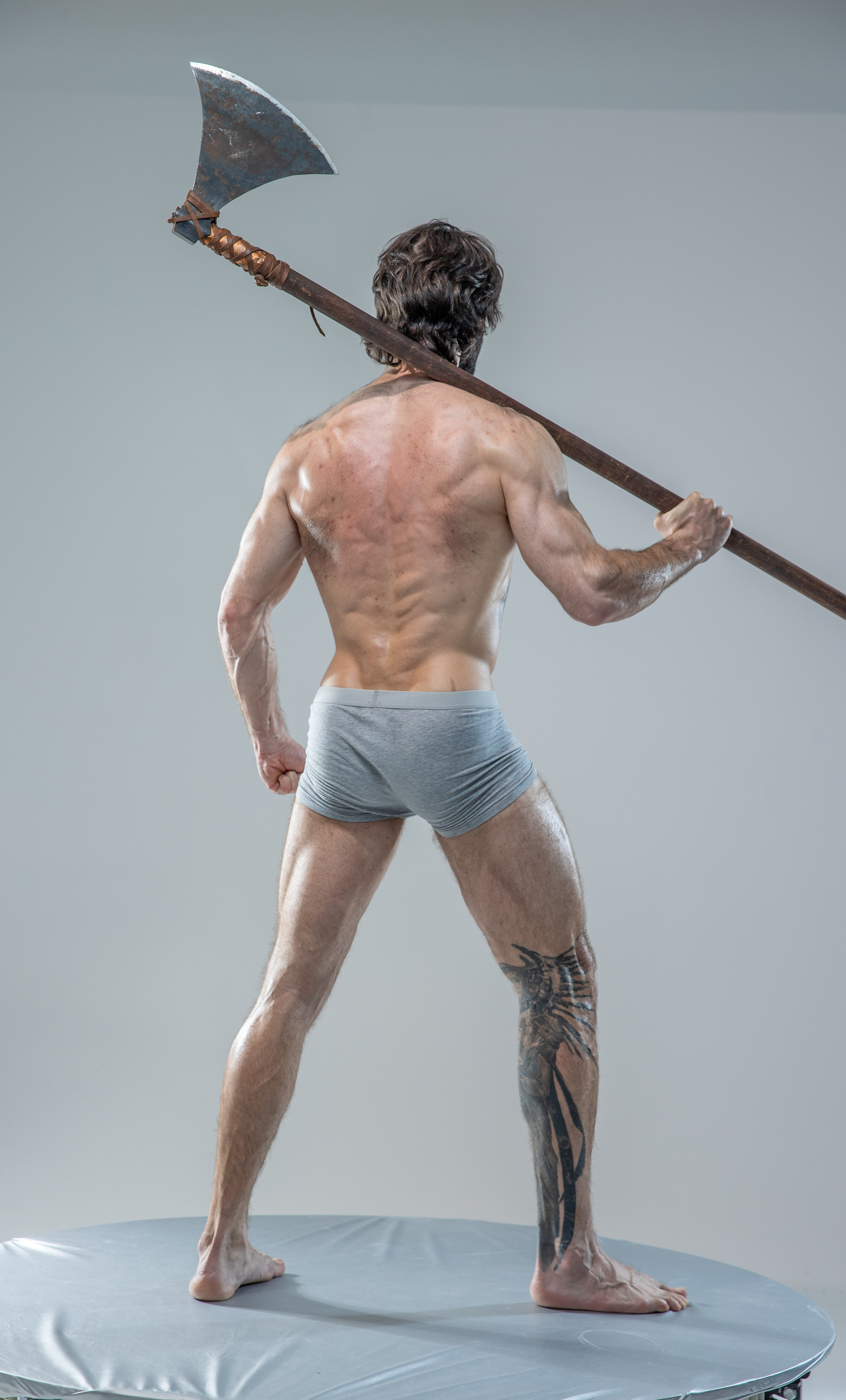 a muscular man stands with his back to the camera, showcasing his toned physique and intricate back tattoo. he holds a large, worn axe over his shoulder,  its handle extending across the image. the neutral studio backdrop and bright artificial lighting emphasize the man's powerful presence.