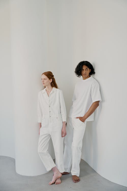 a full shot of a man and a woman standing against a white curved wall. the woman is wearing a white jumpsuit, and the man is wearing a white tshirt and white pants. both are looking away from the camera. the image is minimalist and clean.