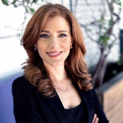 a portrait of a woman in a black suit with long brown hair smiling at the camera. she looks professional, confident, and happy. she's wearing a black jacket and has her arms crossed. there's a plant in the background out of focus.
