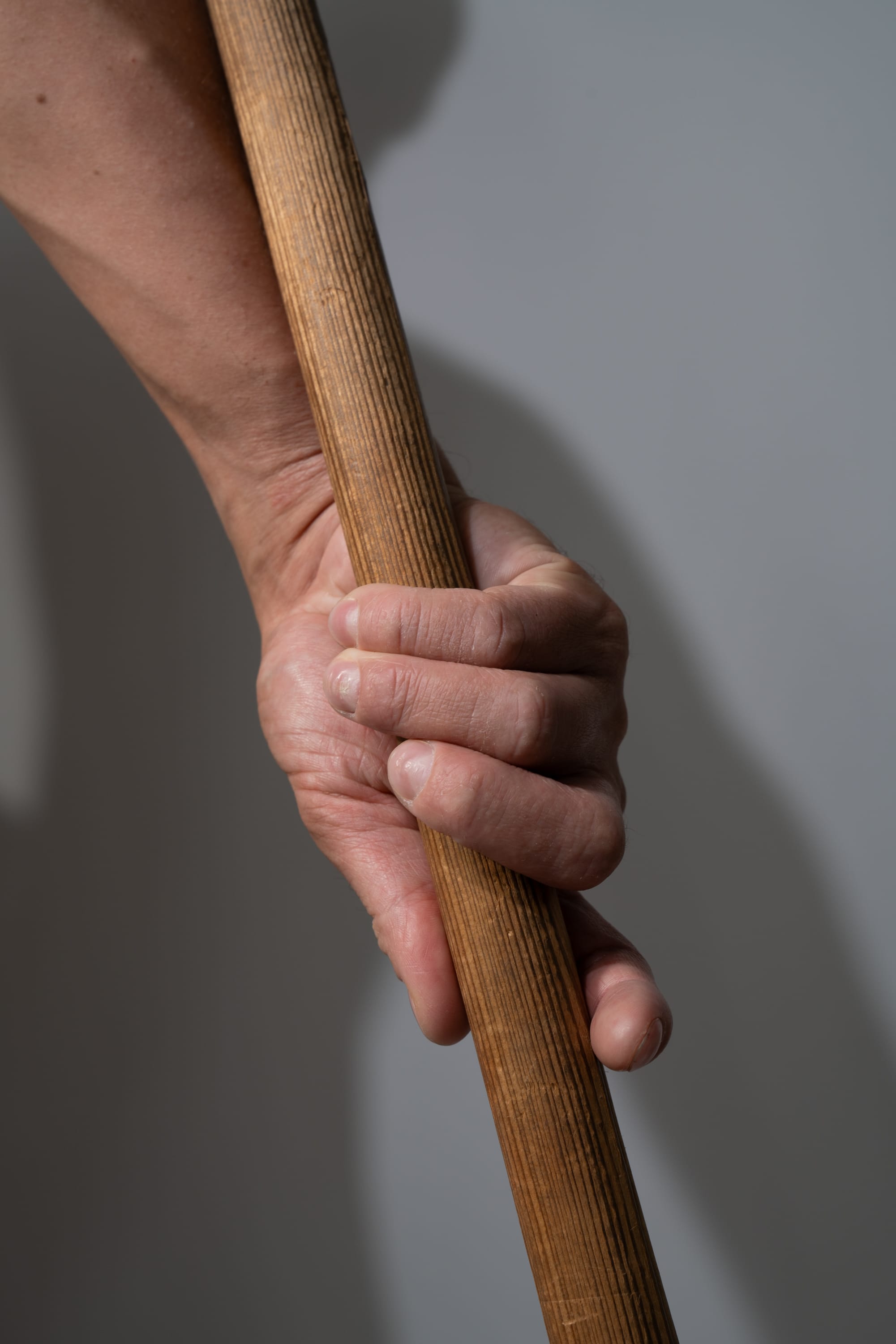 a closeup shot of a man's hand tightly gripping a wooden stick or tool. the hand exhibits signs of age and manual labor, suggesting a life of hard work. the lighting is stark, highlighting the texture of the wood and the veins on the hand. the background is a neutral gray, keeping the focus on the hand and the object.