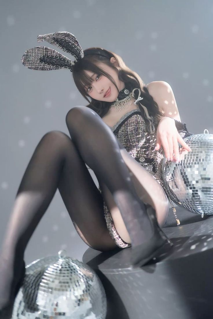 a woman wearing bunny ears and tights is sitting next to disco balls. she is wearing a sparkly dress and a choker. the photo is taken with a grey background and spotlights.
