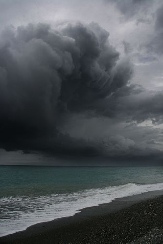 a dramatic seascape shows a turbulent, dark gray storm cloud looming over a sea. the water appears greenishblue, contrasting with the dark sky. a small wave is breaking on a dark pebble beach.