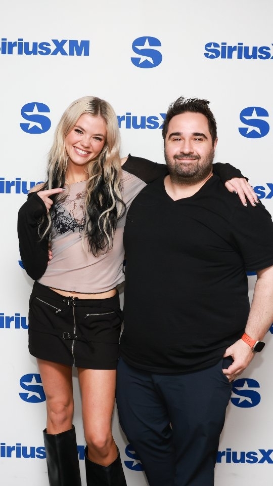 a blonde woman with black streaks in her hair and a darkhaired man are posing together in front of a siriusxm backdrop. the woman is wearing a longsleeve top, a black skirt, and boots. the man is wearing a vneck shirt and dress pants. they are both smiling.