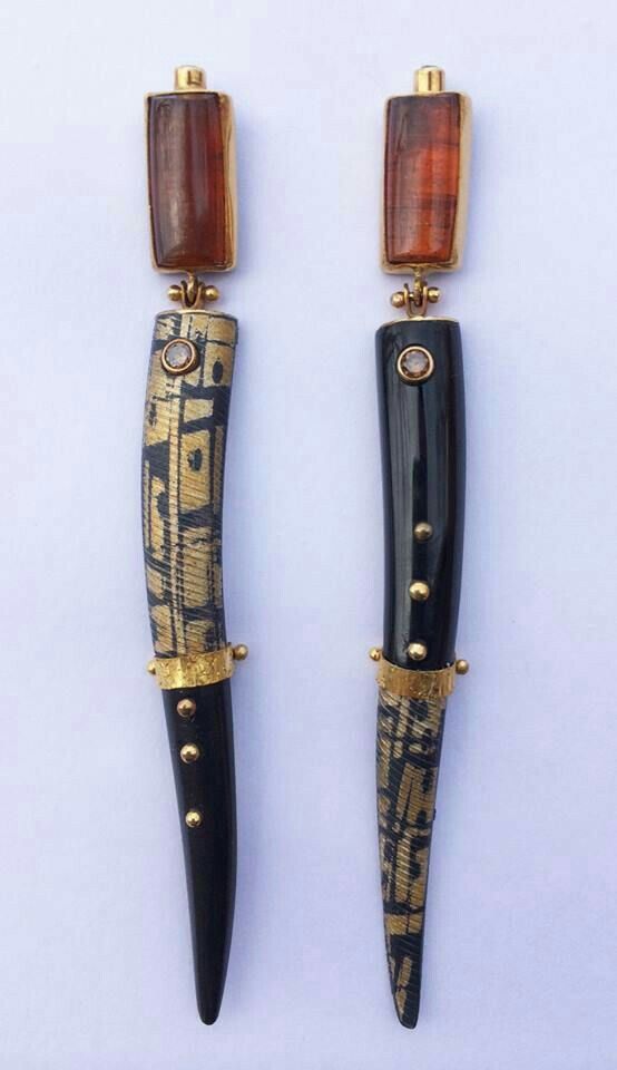 two intricately designed earrings are positioned sidebyside on a light background. each earring features a rectangular brown gemstone at the top, framed in gold. below the gemstone, the earrings transition into elongated, slightly curved hornshaped elements. the left earring has a black horn adorned with a gold pattern and small gold studs, while the right earring has a mostly black horn, also with gold accents and studs. the designs are detailed and visually striking.
