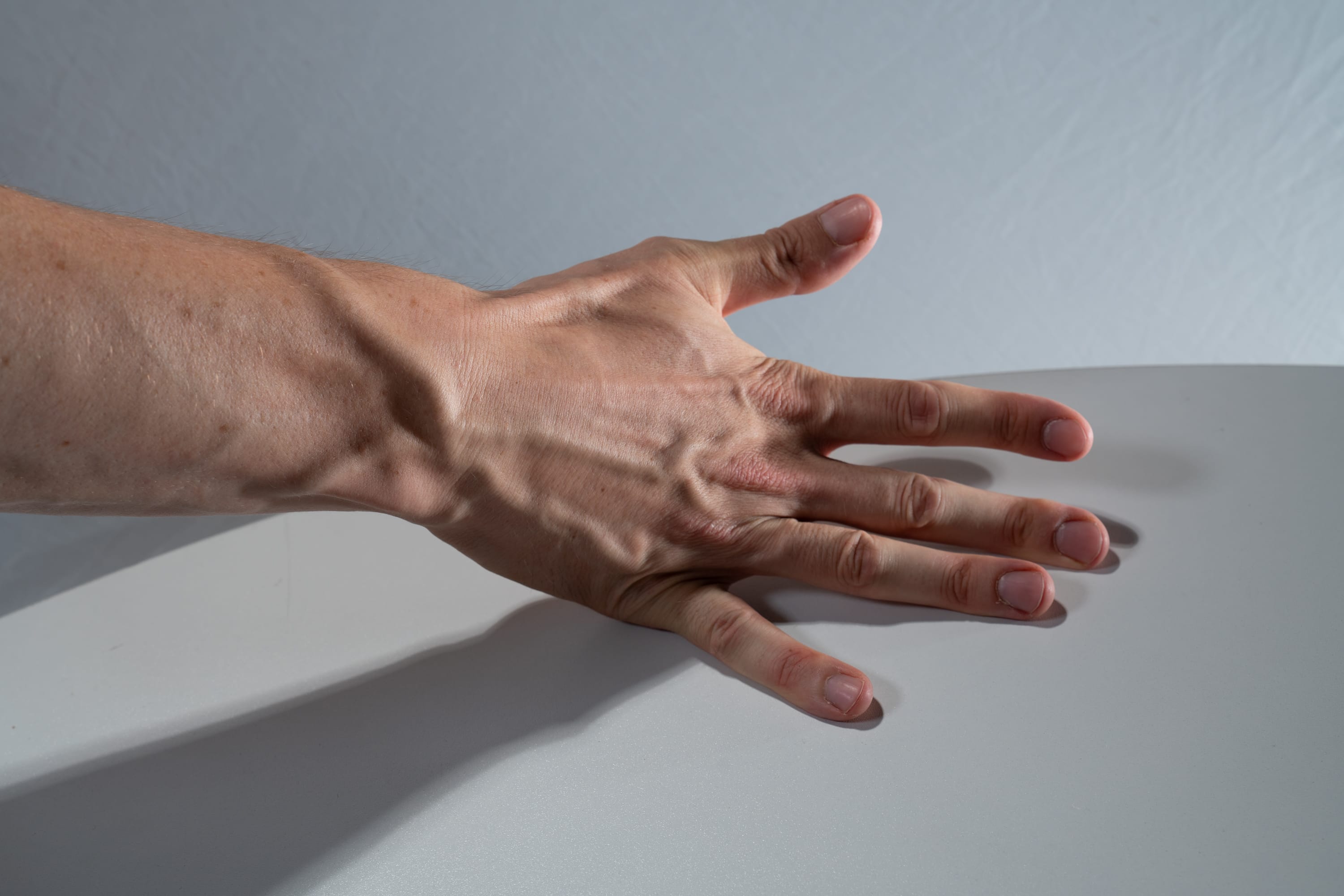the image shows a closeup of a male hand and forearm resting on a smooth, white surface. the hand is outstretched with fingers slightly apart. prominent veins are visible along the back of the hand and forearm due to the directional artificial lighting which creates strong shadows, highlighting the texture and contours of the skin.