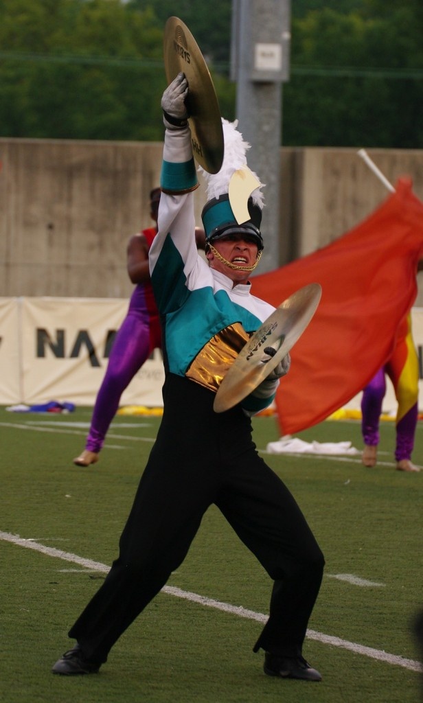a young man in a marching band uniform is playing a cymbal as part of a marching band routine. he is wearing a white and green uniform with a black hat. he is standing on a grassy field. in the background, you can see another band member dressed in purple, and a red flag.