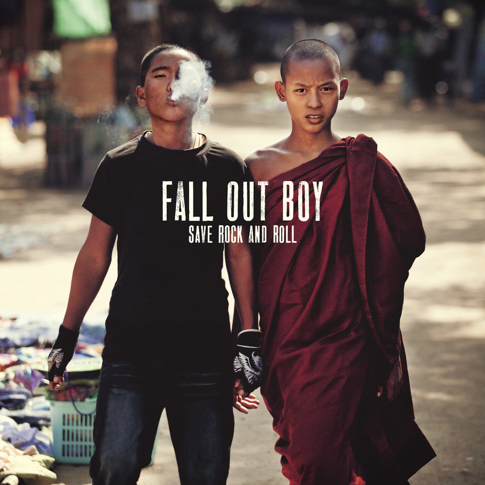 two young men, one wearing black clothing and the other wearing a maroon robe, walk in the middle of a street. the man in the robe is on the right side and has dark hair and his hands are at his side. the man in black is on the left side and has dark hair, his hand is in his pocket. they look like they are looking at the street ahead. both men are young and thin.  in the background there is a street with some buildings and many people in the background. in the center of the image is text that reads "fall out boy save rock and roll" in black text over a white rectangle. the overall image is in focus and is in the style of a rock band album cover.