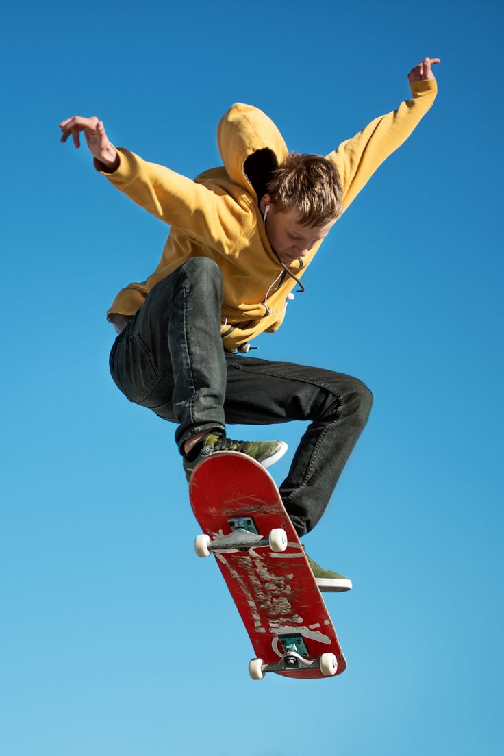 a young skateboarder performs a stunt in the air, with a blue sky as the background. he is wearing a yellow hoodie and black jeans. he is holding his skateboard as he flips through the air. his feet are on the board, and he looks to be having fun as he performs this extreme sport.