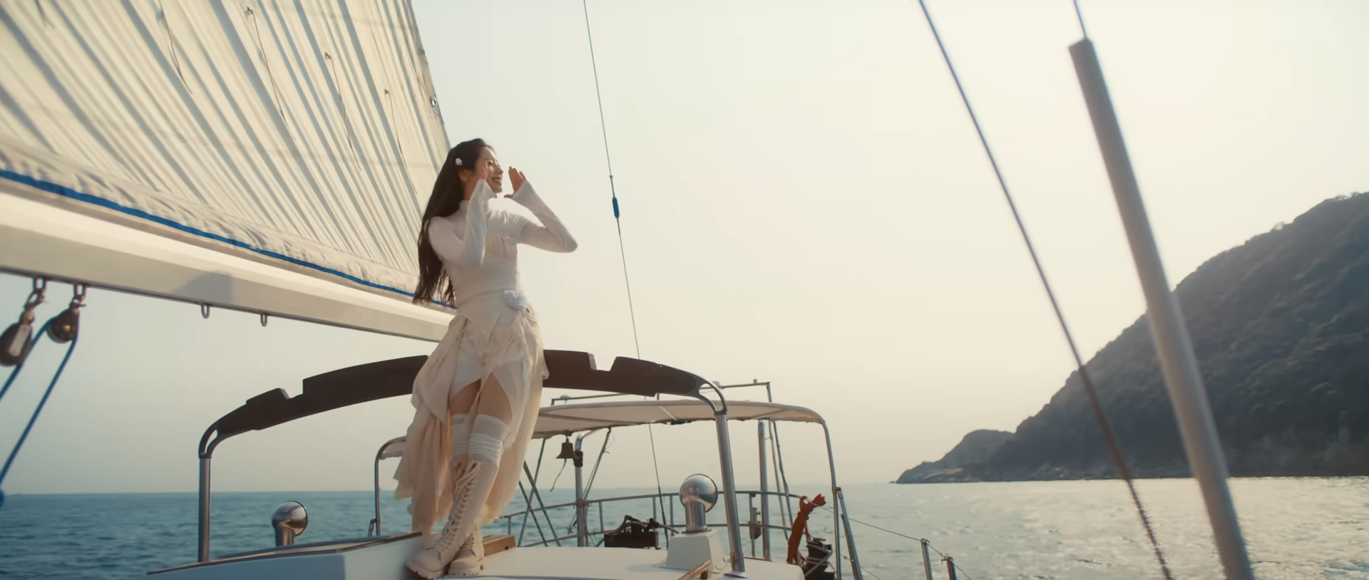 a woman in white clothing stands on the deck of a sailboat, looking out at the ocean and a nearby island. she is wearing a white top, white skirt, and white thighhigh boots. the sails are up, and the boat appears to be moving. the sky is bright and sunny. the ocean is calm and blue. the island is green and mountainous.