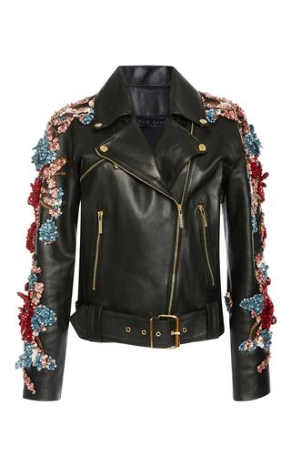 a black leather jacket with floral embroidery on the sleeves. the jacket has a zipper closure, a belt at the waist, and two pockets on the front. the floral embroidery is made of sequins and beads in various colors. the jacket is a biker style with a standup collar and a zipper on the sleeves.