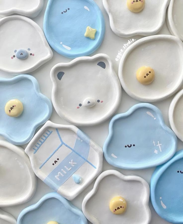 an overhead shot shows a collection of handmade ceramic plates in a variety of cute, cartoonish designs. the plates are decorated with pastel colors and kawaiistyle faces, including a fried egg, a bear, a milk carton, and a cloud. the plates are arranged on a white surface, and they appear to be intended for use as jewelry trays or trinket dishes.