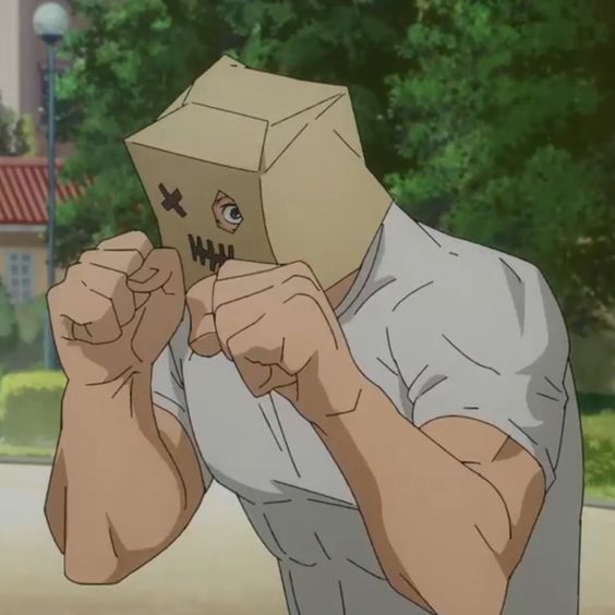 a male anime character with a cardboard box on his head is shown with both fists clenched. he has an angry and determined look on his face. he is outdoors with trees and buildings in the background.  he is dressed in a white tshirt.