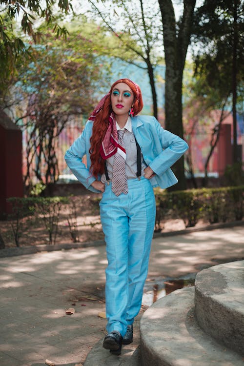 a full shot of a stylish woman in a light blue pantsuit with red hair and a headscarf. she is posing confidently outdoors in a park setting, standing on a stone staircase. her outfit includes a tie and suspenders, adding a retro vibe. the image is welllit, and the woman's makeup is prominent. the overall aesthetic is fashionforward and retroinspired.