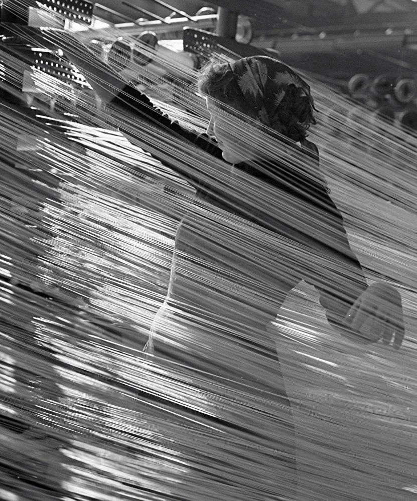 a black and white photo of a woman working in a textile factory. the photo has a strong motion blur, centered on the woman, giving a sense of movement and the busyness of the factory.