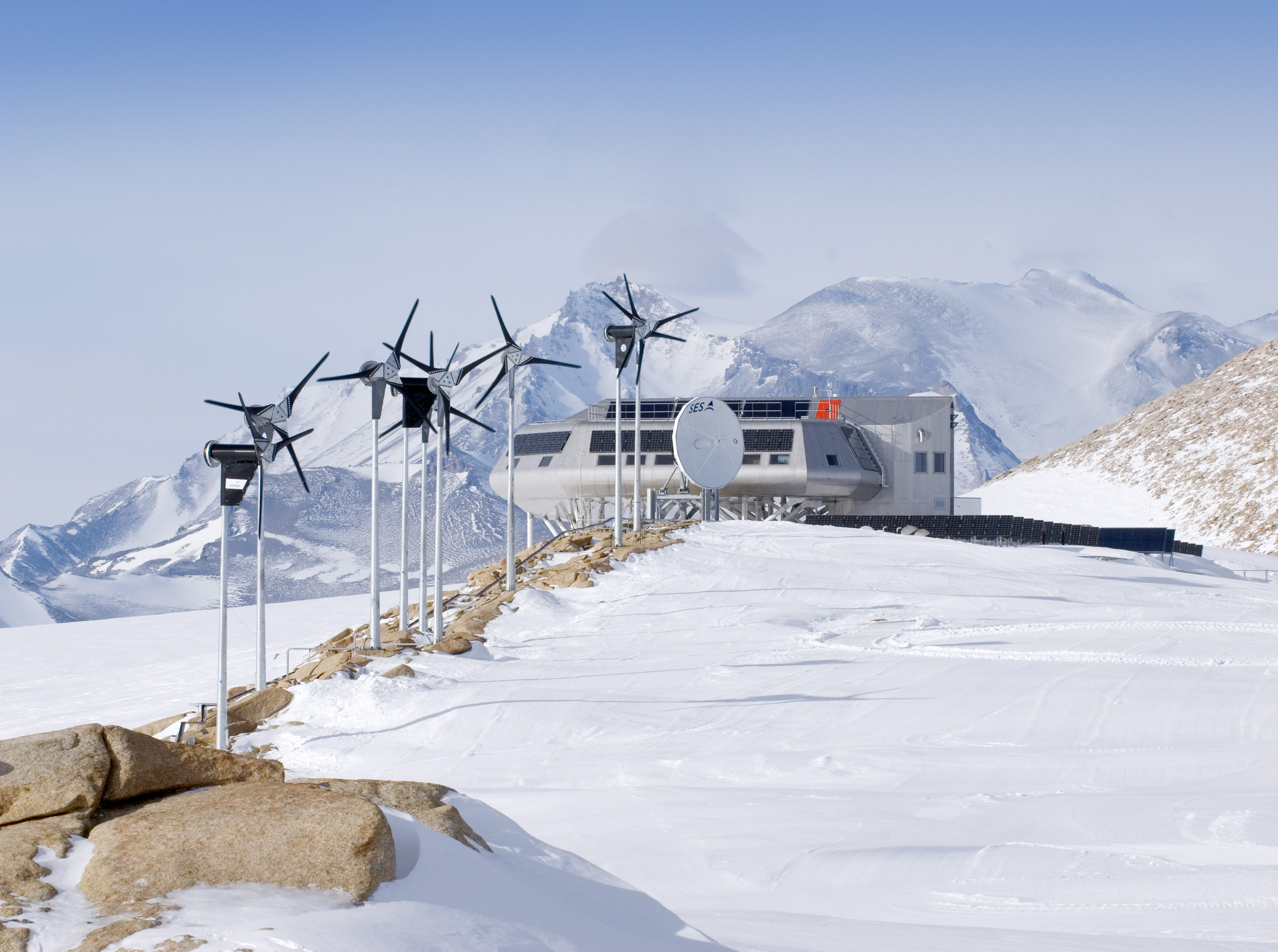 a row of wind turbines stands atop a snowcovered hill overlooking a snowy landscape. behind the turbines is a large, modern building, possibly a research station, nestled among the mountains. the sky is bright blue with a few fluffy clouds, and the ground is covered in a pristine blanket of white. the scene is serene and peaceful, a stark contrast to the cold, harsh environment.  this image suggests the use of renewable energy in a remote, challenging location.