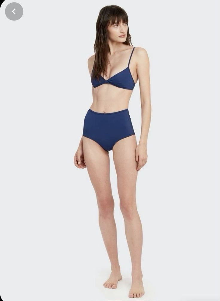 a woman in a blue highwaisted bikini bottom is standing on a white background. she's looking to the side, with her hair slightly covering her face. her arms are at her sides, with her hands slightly in front of her. she is wearing a dark blue bikini top with a black trim and dark blue highwaisted bottoms with a black trim. she is standing in a studio setting.