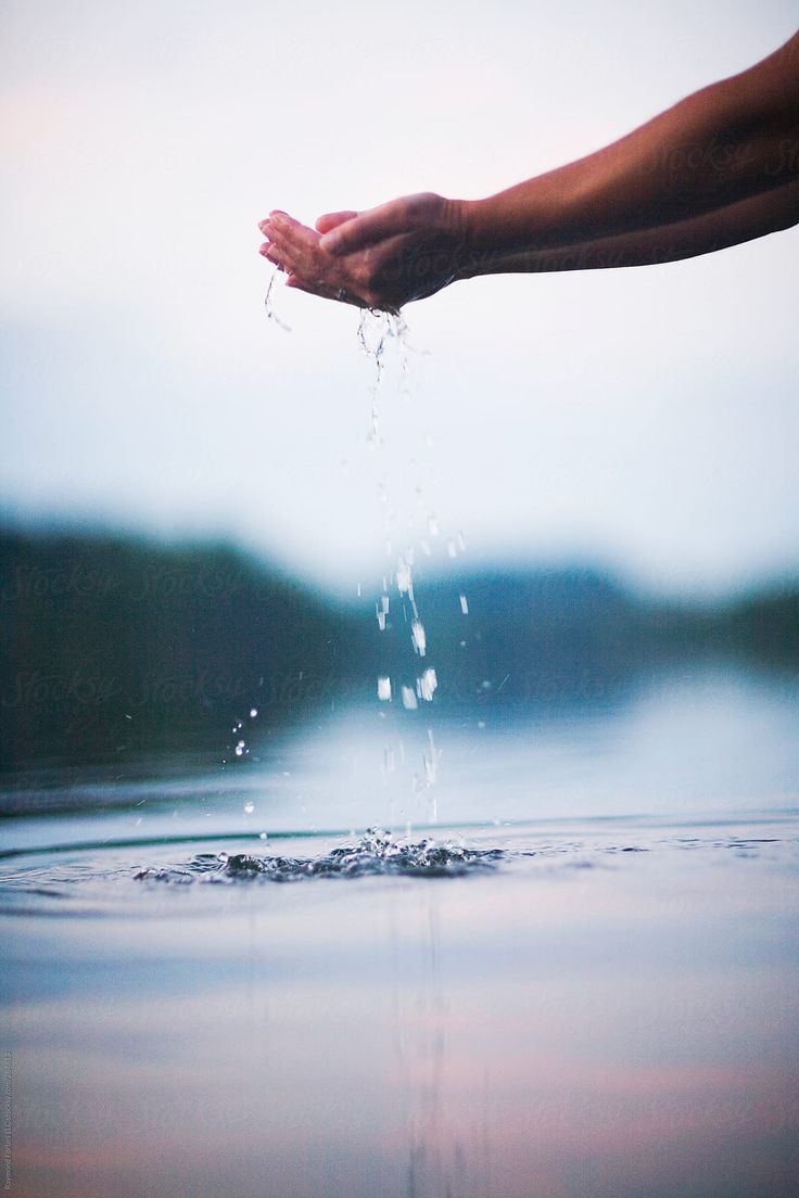 a person's hand is shown cupping water, as the water drops and splashes into a lake. there are soft blue clouds and a gentle mist in the background, giving the image a serene feel.