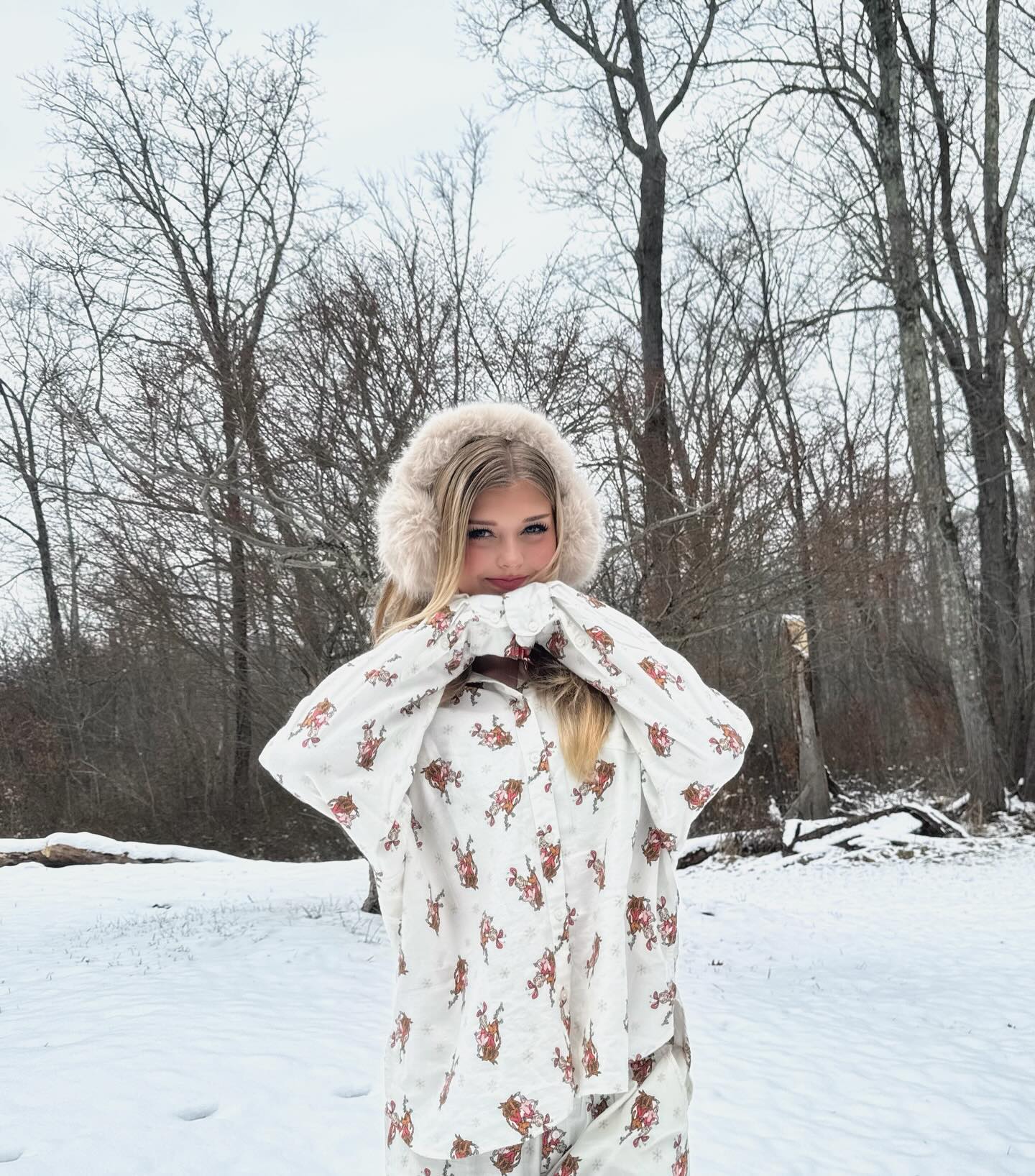 a young woman stands in a snowy landscape wearing pajamas and fur ear muffs. she is smiling and holding her hands up to her face. the background includes snowcovered ground and bare trees.